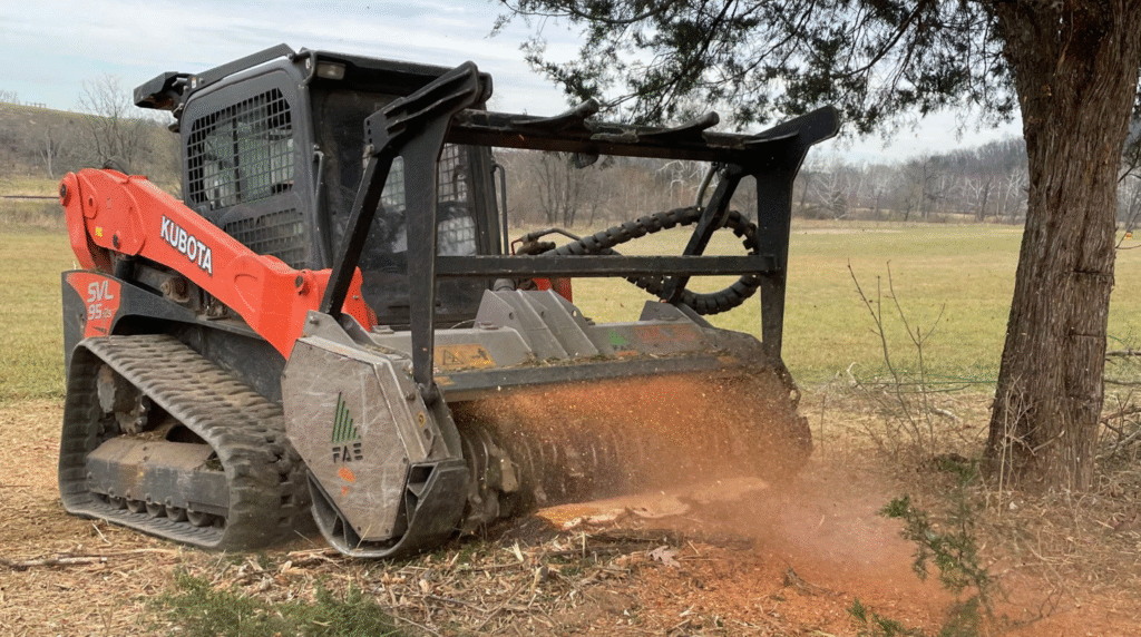 A red and black skid steer loader is actively working on a tree in a construction or landscaping setting.